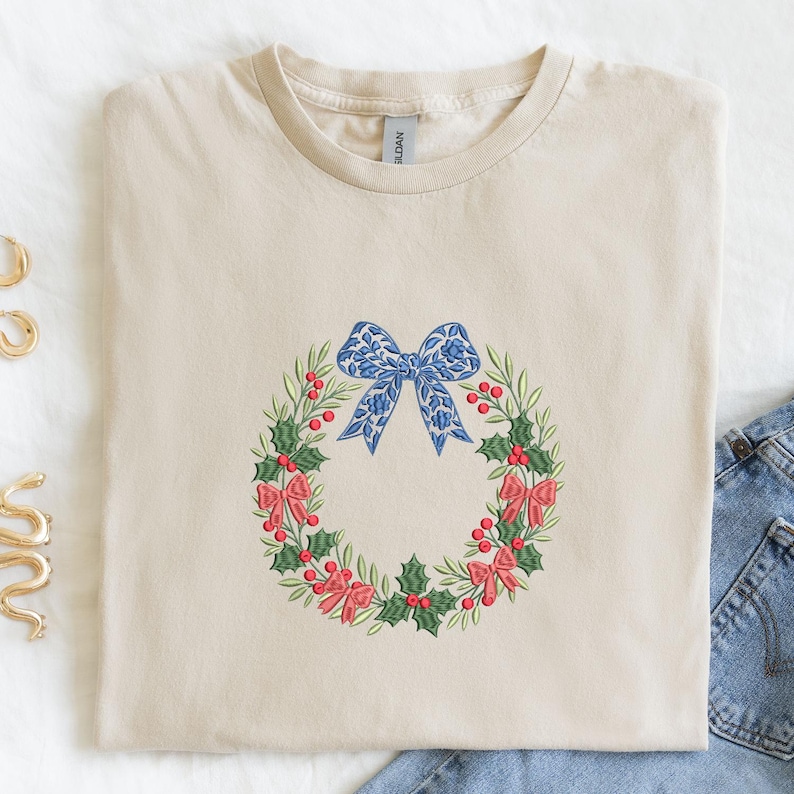 May include: A sand-colored crew neck t-shirt featuring an embroidered Christmas wreath design. The wreath consists of green holly leaves, red berries, and small red bows, topped with a large blue bow featuring a white floral pattern.