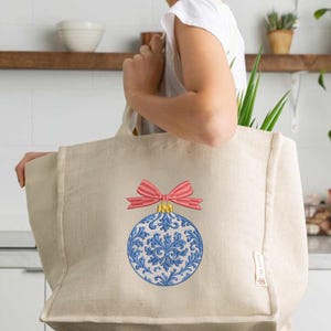May include: A beige canvas tote bag featuring an embroidered design of a round Christmas ornament. The ornament is white with intricate blue floral patterns and is topped with a bright pink bow. The bag has a rectangular shape and a side tag.