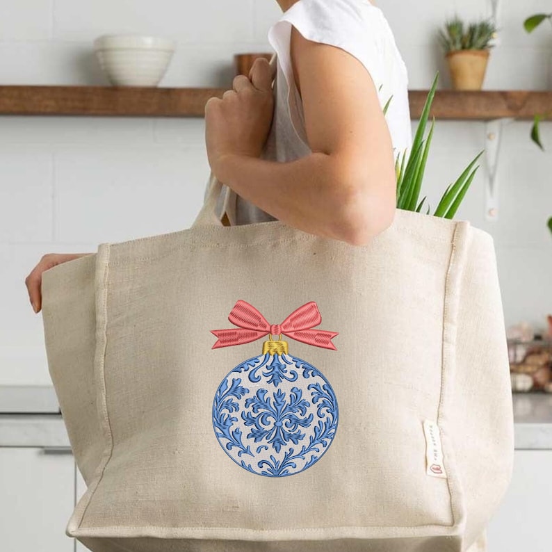 May include: A beige canvas tote bag featuring an embroidered design of a round Christmas ornament. The ornament is white with intricate blue floral patterns and is topped with a bright pink bow. The bag has a rectangular shape and a side tag.