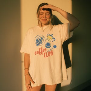 May include: Off-white t-shirt featuring a coffee-themed illustration. The design includes a croissant, coffee pot, cup, and spoon, with the words "coffee club" in red and blue. The shirt is worn by a person.
