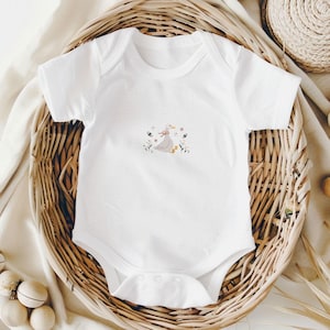 Op de afbeelding: Witte babybody met een geborduurd ontwerp. Het ontwerp toont een grijze gans met een roze strik, omringd door kleurrijke bloemen en vlinders. De body is gemaakt van zacht materiaal en heeft drukknoopsluitingen.
