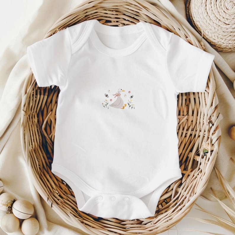 Op de afbeelding: Witte babybody met een geborduurd ontwerp. Het ontwerp toont een grijze gans met een roze strik, omringd door kleurrijke bloemen en vlinders. De body is gemaakt van zacht materiaal en heeft drukknoopsluitingen.