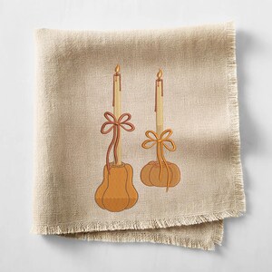 May include: A beige linen napkin with a fringed edge, embroidered with two candles. The candles are atop orange pumpkin-shaped bases and adorned with orange bows. The candle flames are yellow.