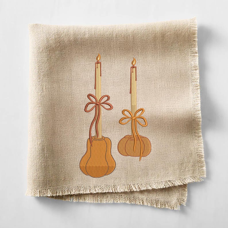 May include: A beige linen napkin with a fringed edge, embroidered with two candles. The candles are atop orange pumpkin-shaped bases and adorned with orange bows. The candle flames are yellow.
