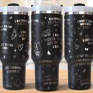 May include: Three black stainless steel travel mugs with silver lettering and butterfly designs. The mugs have different sayings on them, including "I am fucking awesome", "I am fucking perfect just the way I am", and "I don't owe nobody shit".