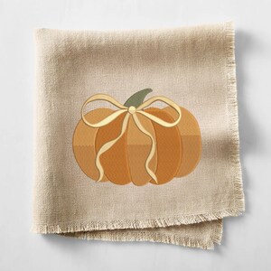 May include: A beige linen napkin with a fringed edge. The napkin features an embroidered pumpkin design in shades of orange and brown, with a cream-colored bow and green stem. A seasonal autumn-themed item.