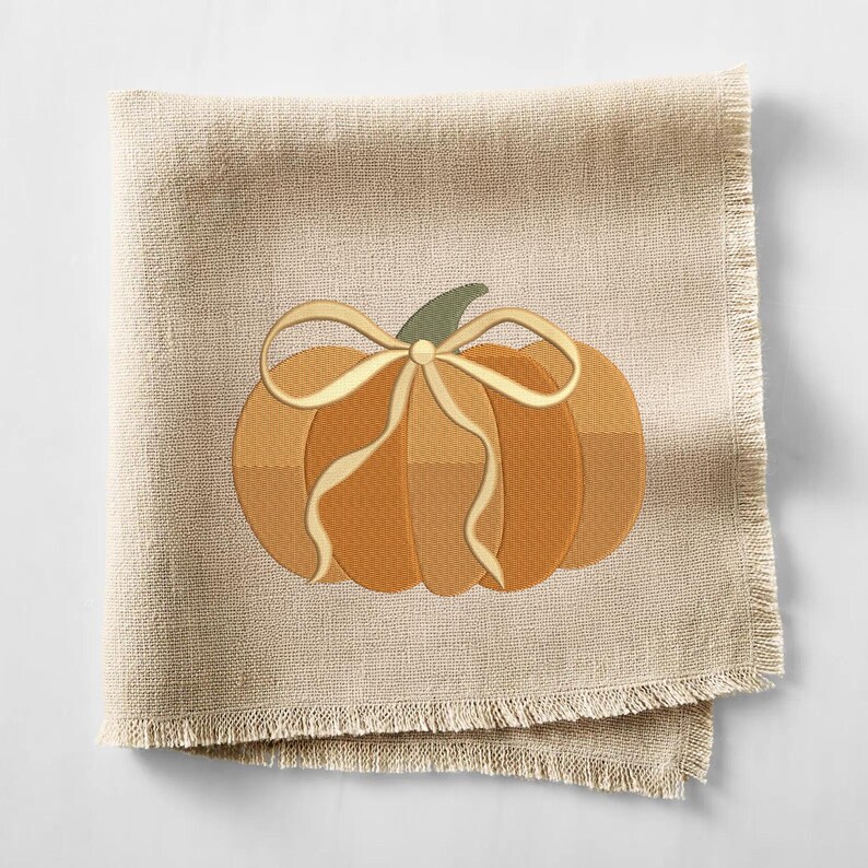 May include: A beige linen napkin with a fringed edge. The napkin features an embroidered pumpkin design in shades of orange and brown, with a cream-colored bow and green stem. A seasonal autumn-themed item.