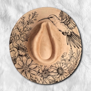 May include: A tan-colored fedora hat with a detailed black ink illustration of a hummingbird and floral designs. The brim features large sunflowers and other flowers. The hat has a teardrop-shaped crown.
