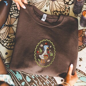 Puede incluir: Una sudadera marr&oacute;n con un dise&ntilde;o bordado de un ganso con un gorro azul y una cesta de fresas. El dise&ntilde;o est&aacute; enmarcado por un borde verde festoneado. La sudadera est&aacute; hecha de un material suave.
