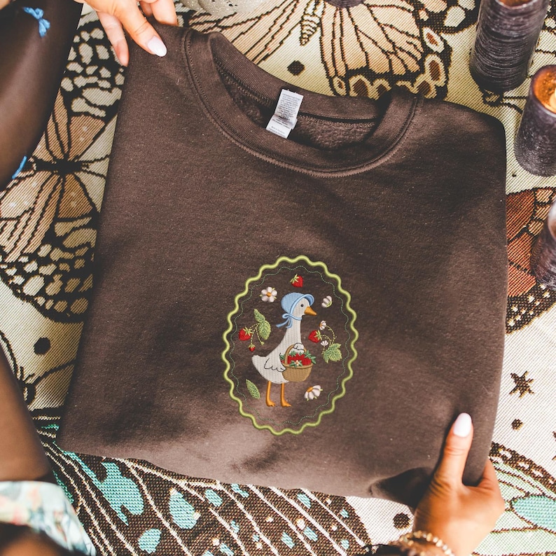 Puede incluir: Una sudadera marr&oacute;n con un dise&ntilde;o bordado de un ganso con un gorro azul y una cesta de fresas. El dise&ntilde;o est&aacute; enmarcado por un borde verde festoneado. La sudadera est&aacute; hecha de un material suave.