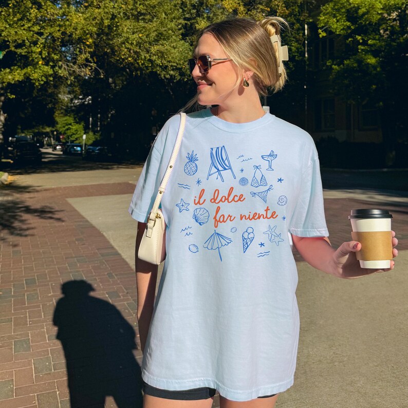 Puede incluir: Una camiseta de manga corta de color azul claro con un estampado gr&aacute;fico en azul y naranja. El dise&ntilde;o incluye peque&ntilde;as ilustraciones de una silla de playa, pi&ntilde;a, concha, helado y bikini, con el texto "il dolce far niente" en color naranja.