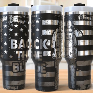 May include: A black and silver stainless steel tumbler with a handle. The tumbler features a distressed American flag design with the words "Back the Blue" and a silhouette of a police officer saluting.