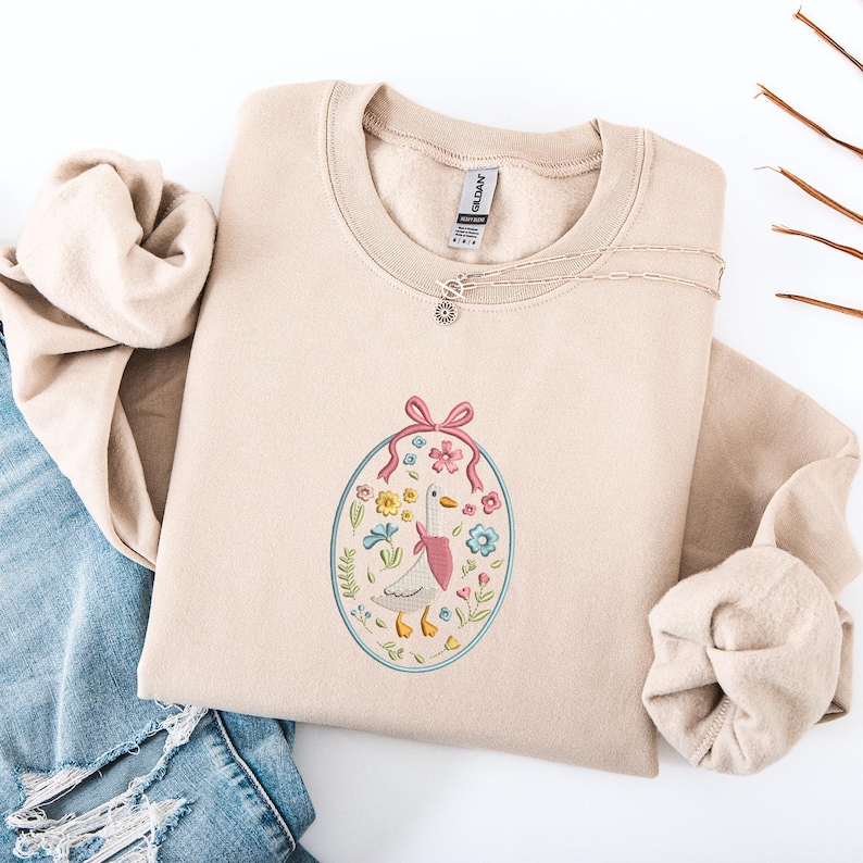 Puede incluir: Sudadera beige con un dise&ntilde;o bordado de un ganso con una bufanda rosa y rodeado de flores. El dise&ntilde;o est&aacute; dentro de un marco ovalado con un lazo rosa en la parte superior. Un collar plateado est&aacute; drapeado sobre la sudadera.