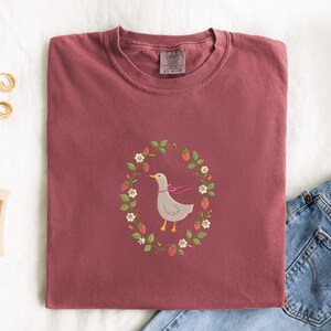 Puede incluir: Camiseta Comfort Colors burdeos con un dise&ntilde;o bordado de un ganso gris con un lazo rosa, rodeado por una corona de fresas y flores. Se ve parte de unos vaqueros.