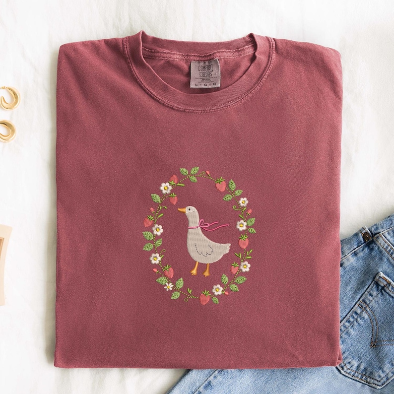 Puede incluir: Camiseta Comfort Colors burdeos con un dise&ntilde;o bordado de un ganso gris con un lazo rosa, rodeado por una corona de fresas y flores. Se ve parte de unos vaqueros.