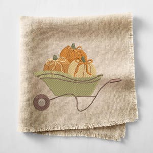 May include: A beige linen napkin with a decorative embroidered design. The design features three pumpkins in a green wheelbarrow. The pumpkins are orange and cream with tan bows. The napkin has a fringed edge.