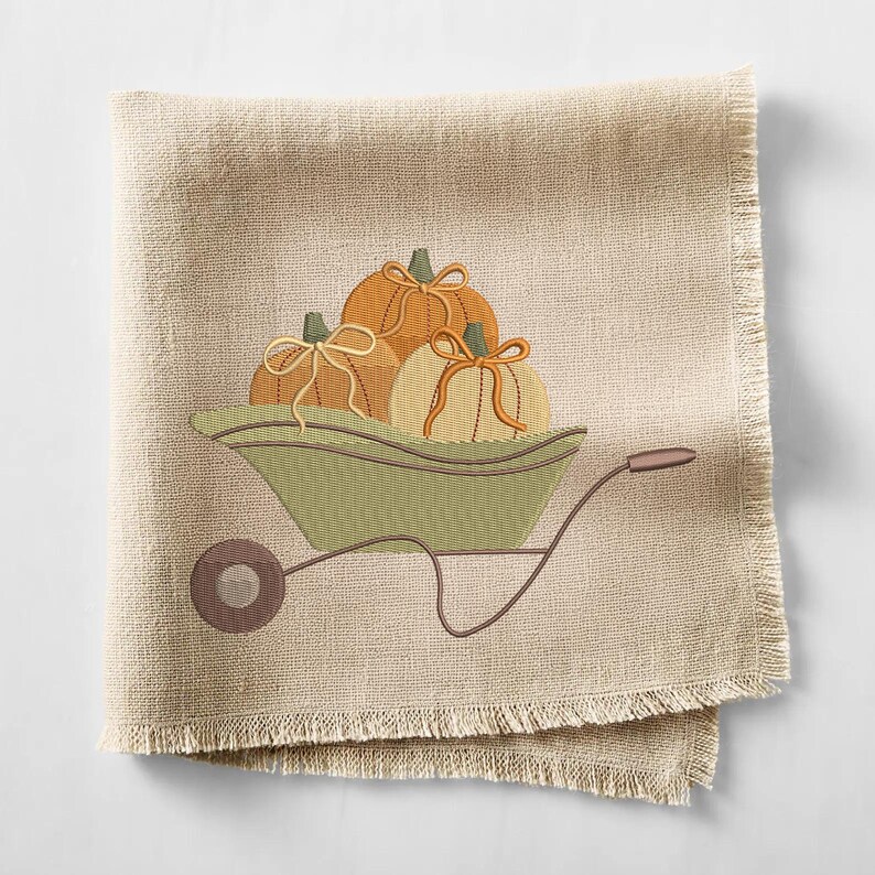 May include: A beige linen napkin with a decorative embroidered design. The design features three pumpkins in a green wheelbarrow. The pumpkins are orange and cream with tan bows. The napkin has a fringed edge.