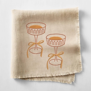 May include: Two embroidered dessert stands with a round top and a bow. The stands are brown with a light brown filling and a yellow bow. The embroidery is on a beige linen napkin with a fringed edge.