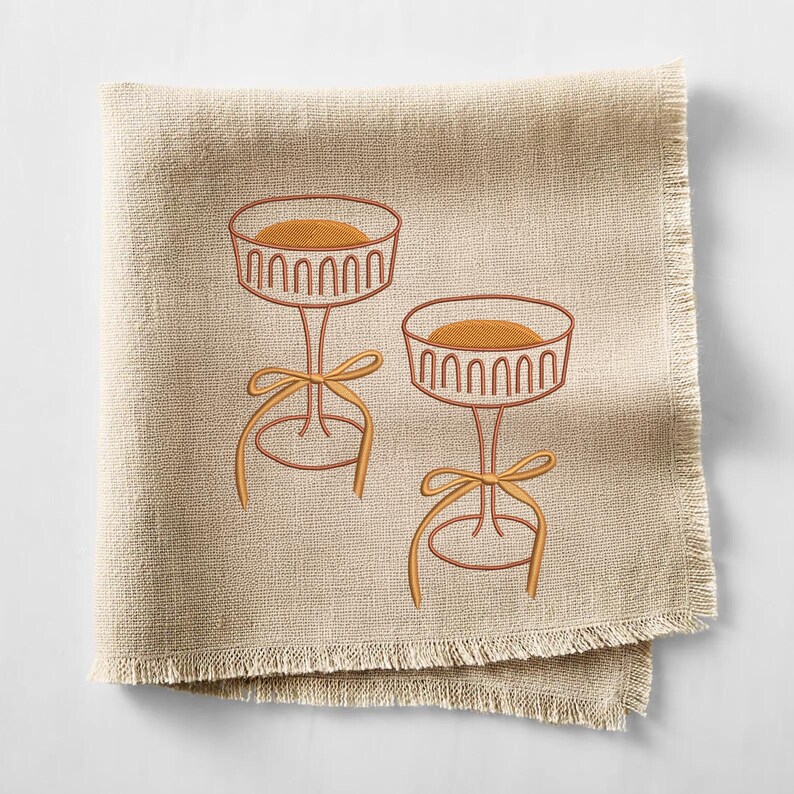 May include: Two embroidered dessert stands with a round top and a bow. The stands are brown with a light brown filling and a yellow bow. The embroidery is on a beige linen napkin with a fringed edge.