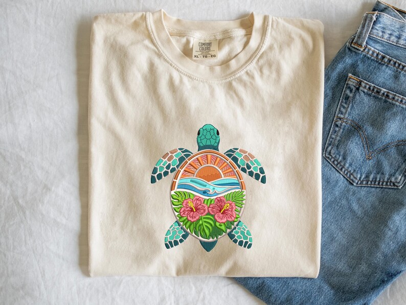 May include: Cream-colored t-shirt featuring a colorful sea turtle design. The turtle has a turquoise head and flippers, with a shell design of orange, blue, and green. The design includes a sunset, waves, and hibiscus flowers.