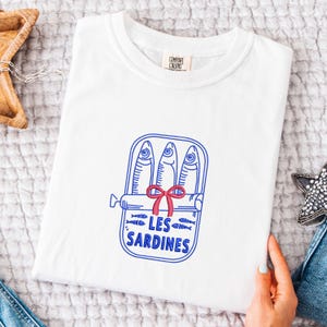 Peut inclure: Un t-shirt blanc &agrave; manches courtes avec un motif graphique en lignes bleues repr&eacute;sentant une bo&icirc;te de sardines contenant trois poissons. Un n&oelig;ud rouge est attach&eacute; au centre de la bo&icirc;te. Le texte LES SARDINES est imprim&eacute; en bleu sous les poissons.