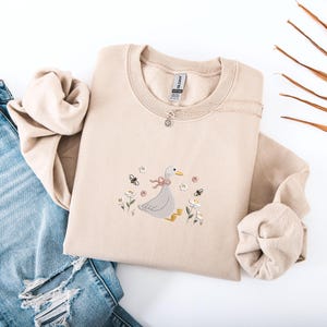 Op de afbeelding: Beige crewneck sweatshirt met een geborduurd ontwerp van een gans met een roze strik, omringd door bloemen en bijen. Een zilveren ketting met een kleine hanger ligt op de sweatshirt. De sweatshirt is opgevouwen op een spijkerbroek.
