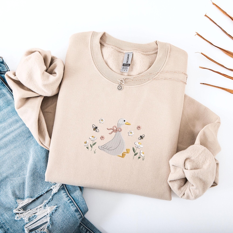 Op de afbeelding: Beige crewneck sweatshirt met een geborduurd ontwerp van een gans met een roze strik, omringd door bloemen en bijen. Een zilveren ketting met een kleine hanger ligt op de sweatshirt. De sweatshirt is opgevouwen op een spijkerbroek.