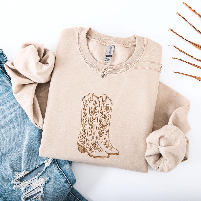 May include: A beige sweatshirt featuring an embroidered design of cowboy boots with floral accents. The boots are in a light brown color and have a detailed stitch pattern. The sweatshirt is folded, with a pair of blue jeans visible in the background.