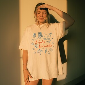 Puede incluir: Una camiseta de manga corta color blanco roto con el texto rojo "il dolce far niente" centrado en el pecho. El dise&ntilde;o est&aacute; rodeado de ilustraciones de l&iacute;neas azules de una pi&ntilde;a, una silla de playa, un bikini, una concha, una sombrilla, un helado y una estrella de mar.