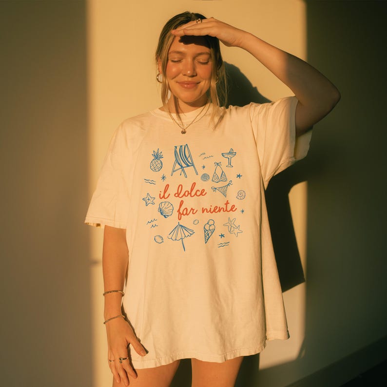Puede incluir: Una camiseta de manga corta color blanco roto con el texto rojo "il dolce far niente" centrado en el pecho. El dise&ntilde;o est&aacute; rodeado de ilustraciones de l&iacute;neas azules de una pi&ntilde;a, una silla de playa, un bikini, una concha, una sombrilla, un helado y una estrella de mar.