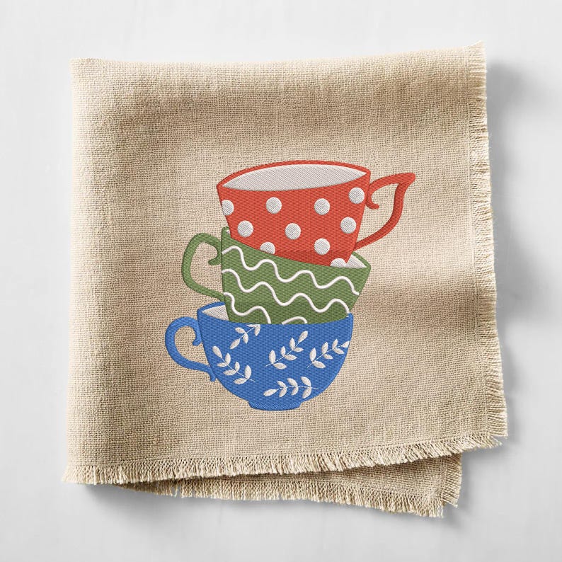 May include: A beige linen napkin with a stack of three embroidered teacups. The top cup is red with white polka dots, the middle is green with a wavy pattern, and the bottom is blue with white leaf designs. The napkin has a fringed edge.