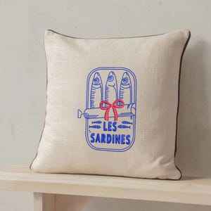 Peut inclure: Un coussin carr&eacute; beige avec un passepoil marron fonc&eacute;. Le centre pr&eacute;sente un dessin au trait bleu repr&eacute;sentant une bo&icirc;te de sardines avec trois poissons, un n&oelig;ud rouge et le texte LES SARDINES imprim&eacute; en lettres capitales bleues.