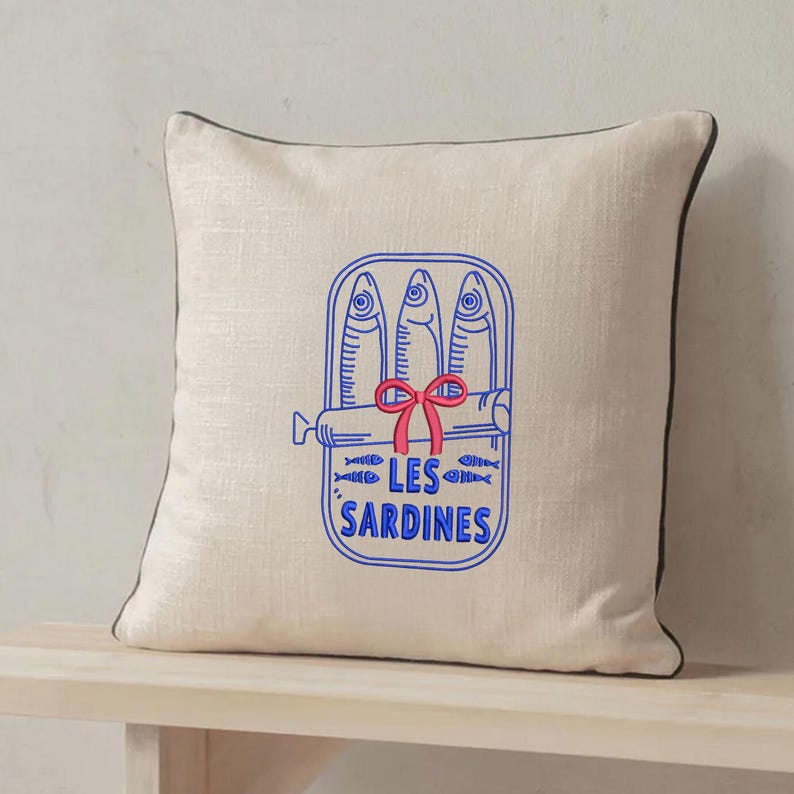 Peut inclure: Un coussin carr&eacute; beige avec un passepoil marron fonc&eacute;. Le centre pr&eacute;sente un dessin au trait bleu repr&eacute;sentant une bo&icirc;te de sardines avec trois poissons, un n&oelig;ud rouge et le texte LES SARDINES imprim&eacute; en lettres capitales bleues.