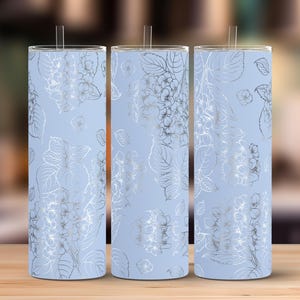 Könnte beinhalten: Drei hellblaue Isolierbecher mit floralem Design. Das Design zeigt silberfarbene Hortensienblüten und -blätter. Jeder Becher hat einen durchsichtigen Strohhalm und einen silberfarbenen Rand.