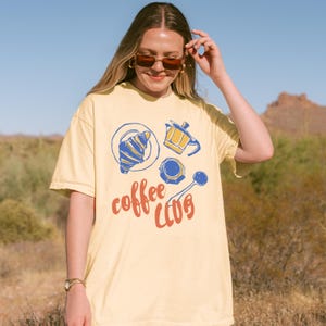 May include: A pale yellow t-shirt featuring a coffee-themed illustration. The design includes a croissant, coffee pot, cup, and spoon, with the words "coffee club" in red script. The shirt is worn by a person wearing sunglasses.