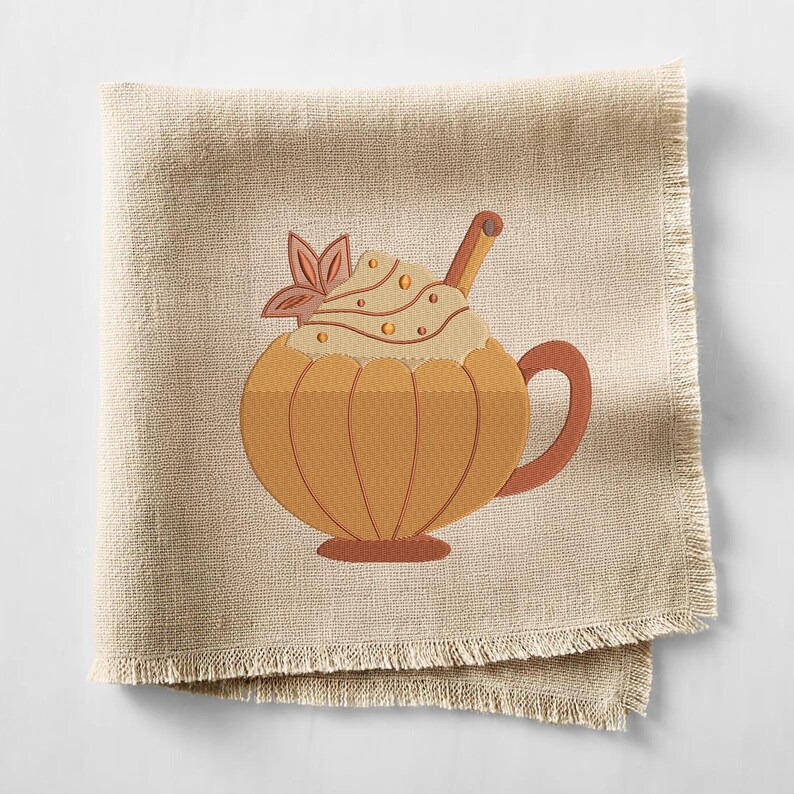 May include: A beige linen napkin with a pumpkin spice latte design. The embroidered latte features a pumpkin-shaped cup, whipped cream, a cinnamon stick, and a decorative leaf. The napkin has a fringed edge.
