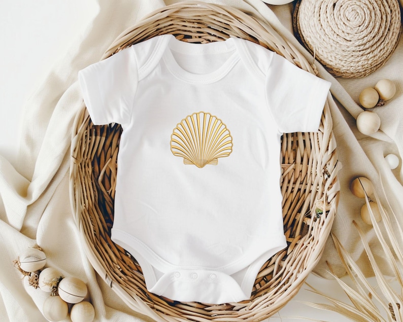 May include: White baby onesie with a gold seashell design. The short-sleeved bodysuit is made of soft material and features a snap closure. The seashell is centered on the front of the onesie. The onesie is displayed in a woven basket.