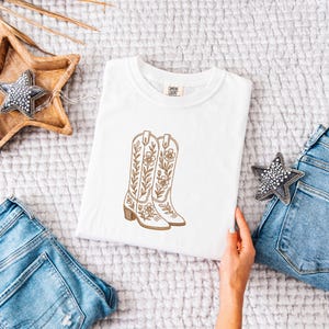 May include: White t-shirt featuring an embroidered design of cowboy boots with floral and leaf details. The boots are in shades of brown and tan. The shirt is displayed on a soft, textured surface, with denim jeans and decorative stars in the background.