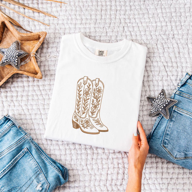 May include: White t-shirt featuring an embroidered design of cowboy boots with floral and leaf details. The boots are in shades of brown and tan. The shirt is displayed on a soft, textured surface, with denim jeans and decorative stars in the background.