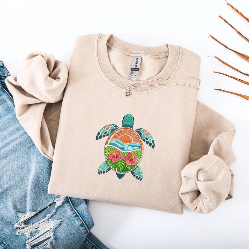 May include: A beige sweatshirt featuring a colorful turtle design. The turtle has a teal head and patterned flippers, with a sunset and ocean scene on its shell. Pink hibiscus flowers and waves are also featured in the design. A silver necklace is draped on the sweatshirt.