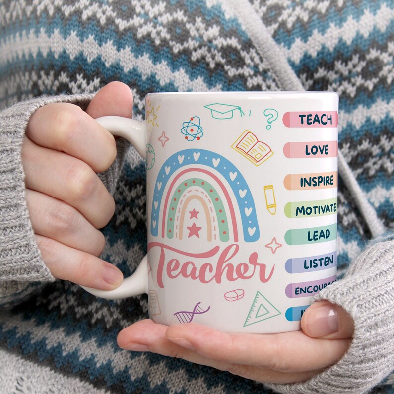 May include: White ceramic mug with a rainbow design and the word "teacher" in pink lettering. The mug has a colorful list of verbs that describe the role of a teacher, including "teach", "love", "inspire", "motivate", "lead", "listen", and "encourage".