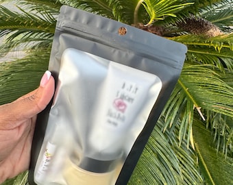 May include: A black resealable pouch with a clear front panel containing a white tube with pink lettering and a small jar with a gold lid. The pouch is hanging from a palm tree.