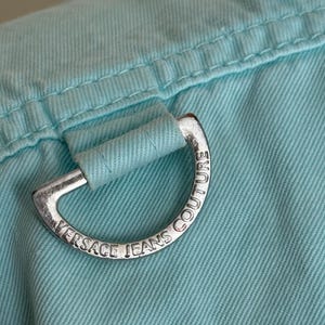 Może przedstawiać: Zbliżenie jasnoniebieskiej tkaniny ze srebrnym metalowym pierścieniem D. Pierścień jest grawerowany napisem "VERSACE JEANS COUTURE". Tkanina ma widoczne szwy i teksturowany wygląd.