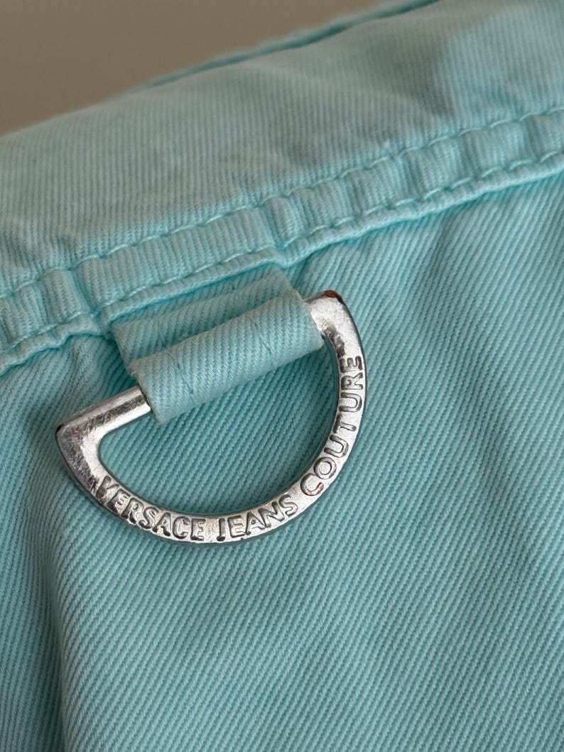 Może przedstawiać: Zbliżenie jasnoniebieskiej tkaniny ze srebrnym metalowym pierścieniem D. Pierścień jest grawerowany napisem "VERSACE JEANS COUTURE". Tkanina ma widoczne szwy i teksturowany wygląd.