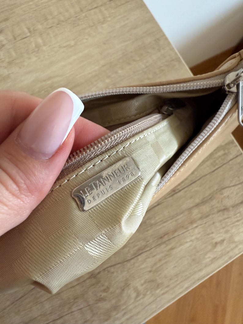 May include: A tan leather handbag with a zipper closure. The interior is lined with a patterned fabric. A metal tag inside the bag reads "LE TANNEUR DEPUIS 1898".