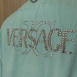 Może przedstawiać: Jasnoniebieska kurtka dżinsowa z napisem "VERSACE" wysadzanym srebrnymi kryształkami na plecach. Kurtka ma srebrną metalową pętlę przy kołnierzu i klasyczny dżinsowy design.
