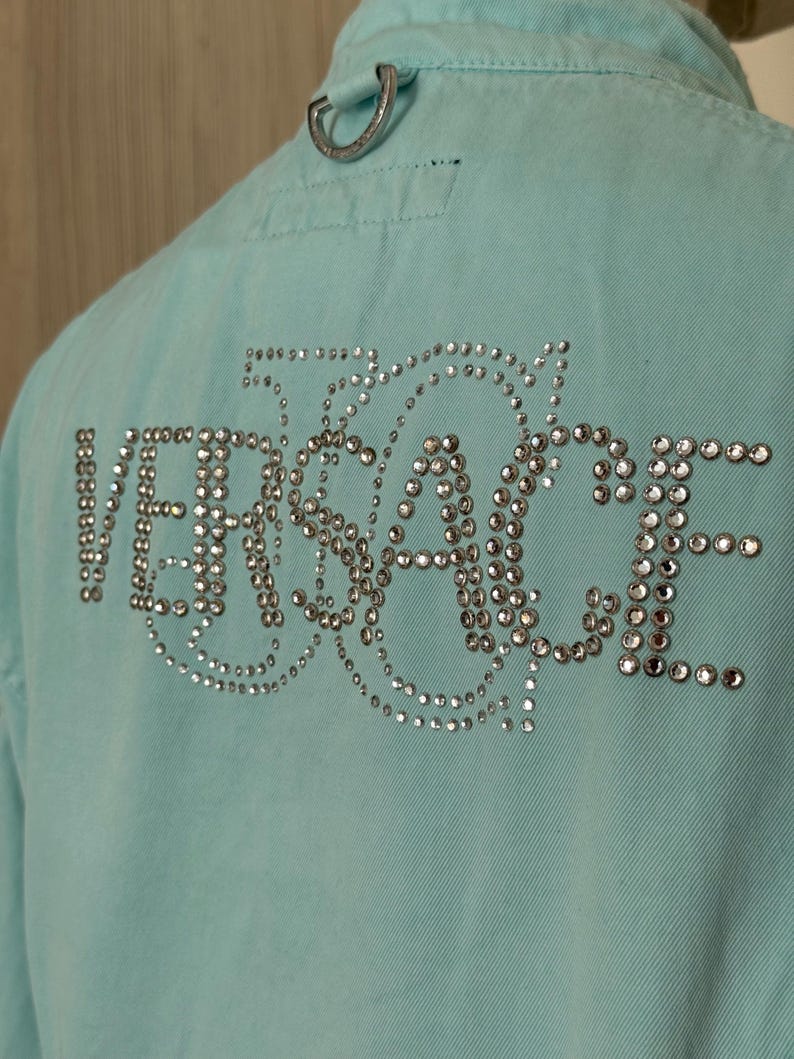 Może przedstawiać: Jasnoniebieska kurtka dżinsowa z napisem "VERSACE" wysadzanym srebrnymi kryształkami na plecach. Kurtka ma srebrną metalową pętlę przy kołnierzu i klasyczny dżinsowy design.