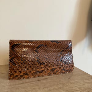 May include: A brown and black snakeskin clutch purse. The rectangular purse has a flap closure and is made of a snakeskin pattern. The purse is sitting on a wooden surface.