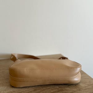 May include: A tan-colored shoulder bag with a long strap. The bag is made of a smooth material and has a rectangular shape. The bag is sitting on a wooden surface.