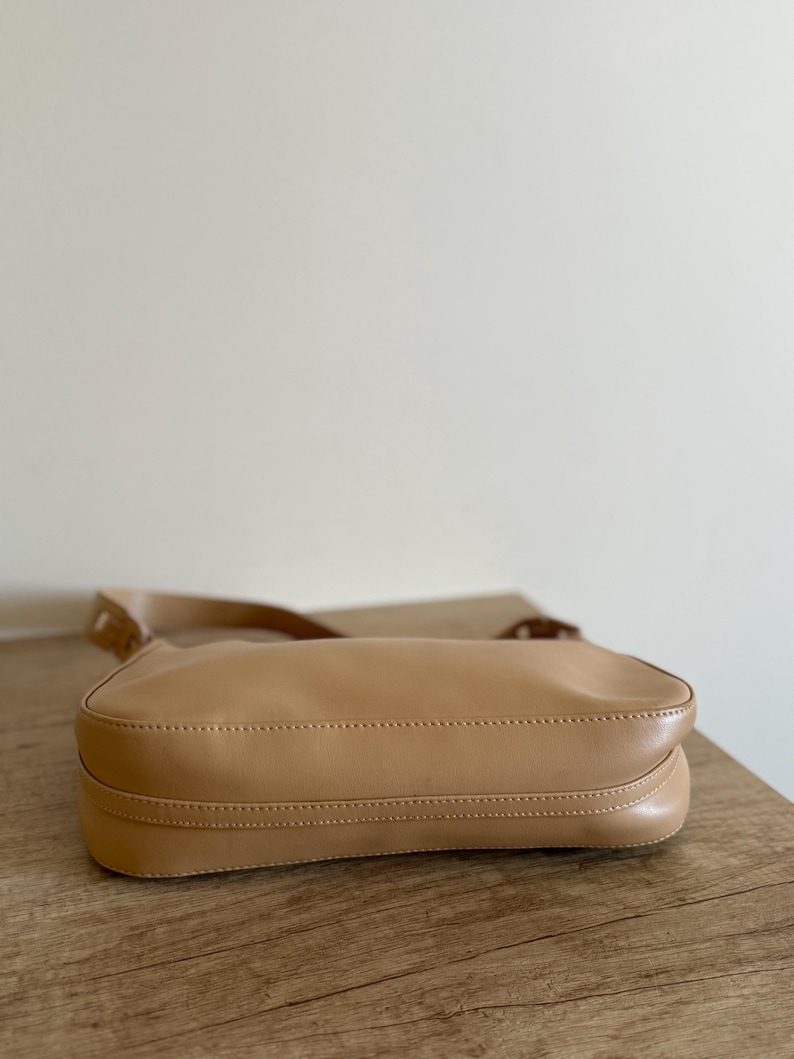 May include: A tan-colored shoulder bag with a long strap. The bag is made of a smooth material and has a rectangular shape. The bag is sitting on a wooden surface.